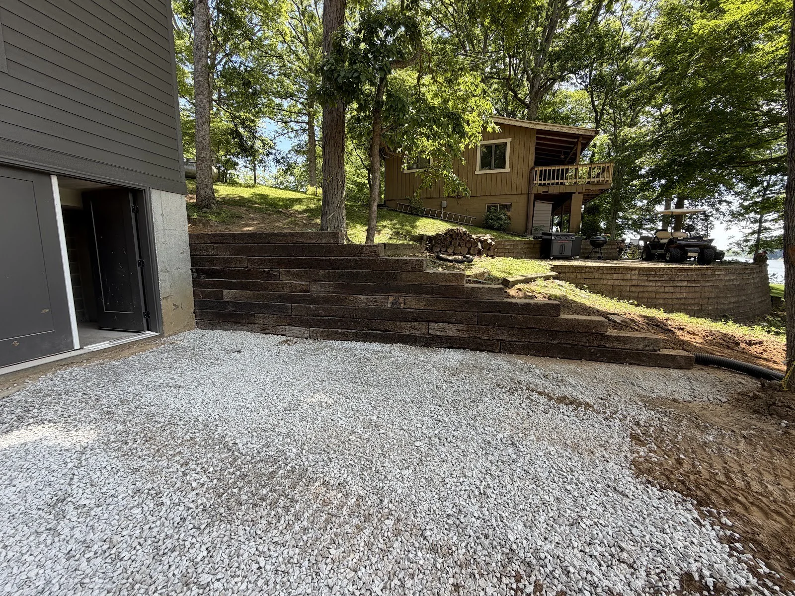 Tiered railroad-tie retaining wall and fresh gravel pad built into a wooded hillside below a lake cottage.